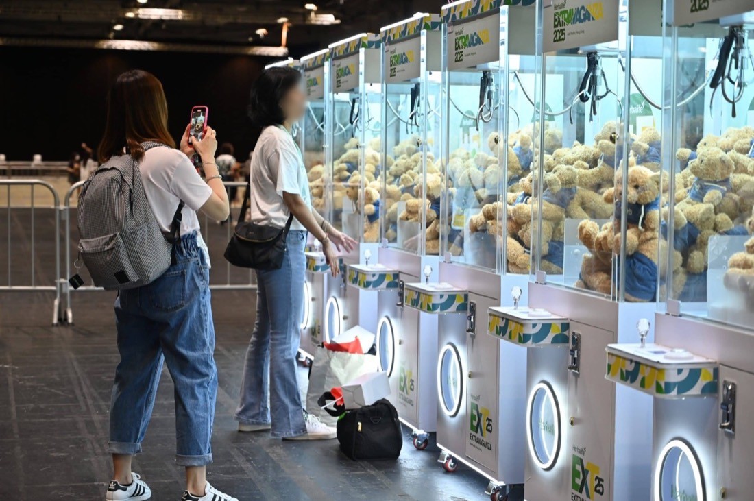 商場活動現場一排品牌夾公仔機,每部機內堆滿同一款穿藍色上衣的米色毛公仔,兩名參觀者在機前拍照