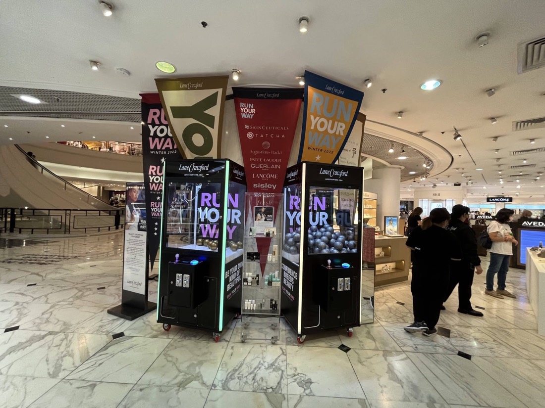 商場中庭擺放兩部黑色機身、藍色燈光的品牌夾公仔機,機頂有品牌標誌,旁邊是橫額與路過的顧客